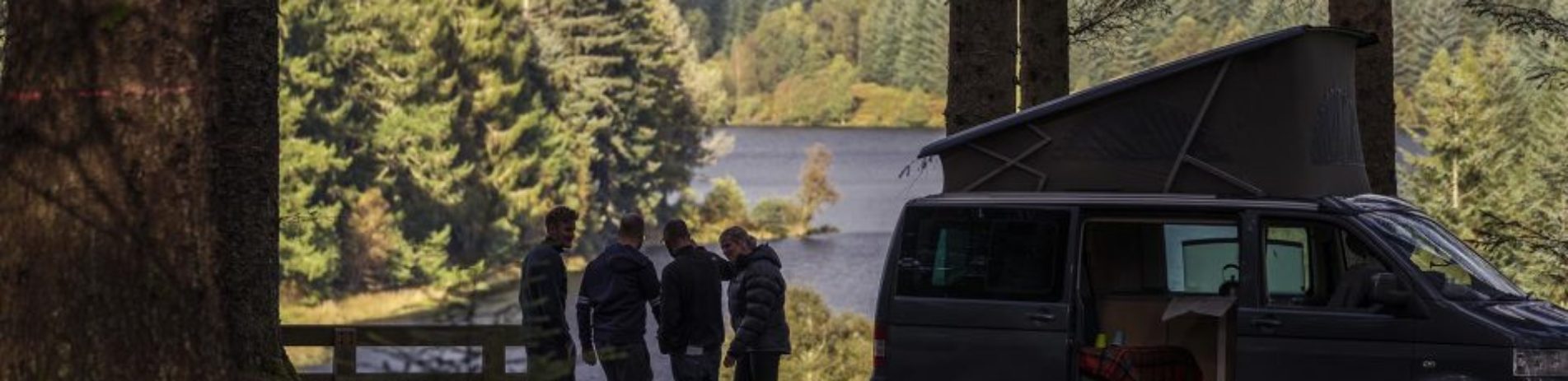 four-friends-chatting-outside-campervan-with-door-open-with-stunning-vista-lit-by-sun-behind-coniferous-forests-and-loch