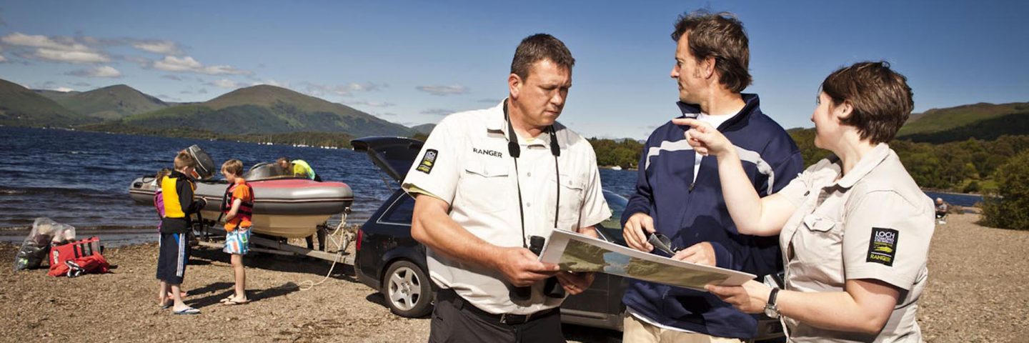 two-rangers-chatting-to-member-of-public-and-pointing-on-map-and-in-the-distance-with-boat-being-launched-in-loch-lomond-next-to-car-and-group-of-children