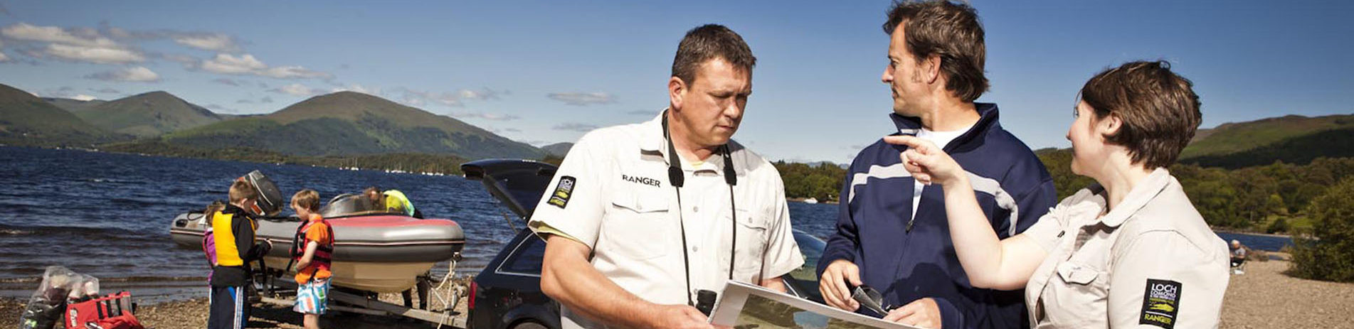 two-rangers-chatting-to-member-of-public-and-pointing-on-map-and-in-the-distance-with-boat-being-launched-in-loch-lomond-next-to-car-and-group-of-children