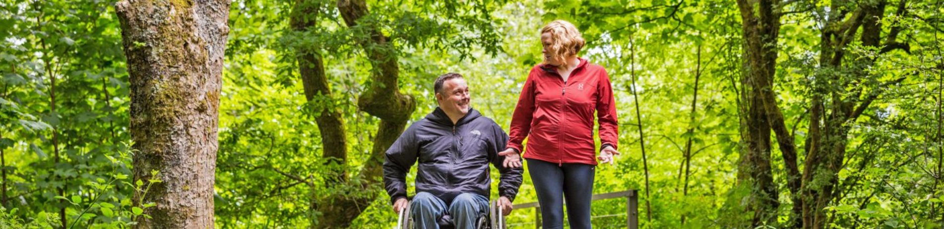 man-in-wheelchair-jeans-and-dark-jacket-accompanied-by-woman-in-red-sports-top-on-park-path-forest-canopy-behind