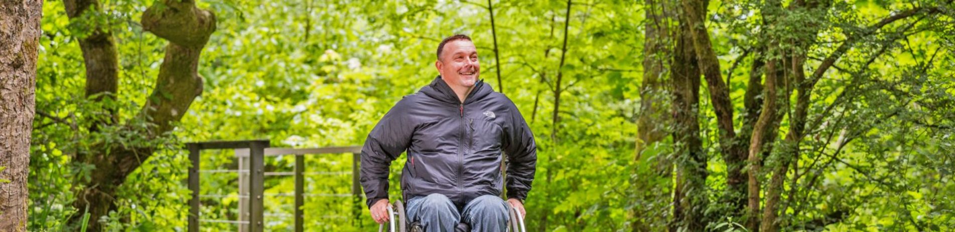 man-in-wheelchair-in-grey-jacket-and-jeans-smiling-surrounded-by-lush-forest-canopy