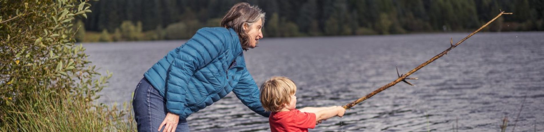 woman-in-blue-outdoor-coat-helping-young-boy-in-red-t-shirt-hold-longh-stick-over-loch-drunkie-mock-fishing