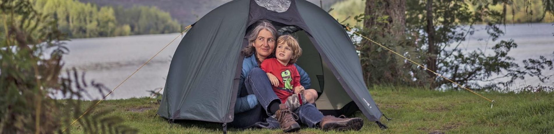 woman-holding-young-boy-at-entrance-of-dark-green-tent-in-the-forest-with-loch-water-visible-behind