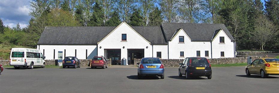 balmaha-visitor-centre-long-white-building-with-grey-slate-roof-and-with-trees-towering-behind-and-an-extensive-car-park-in-the-front