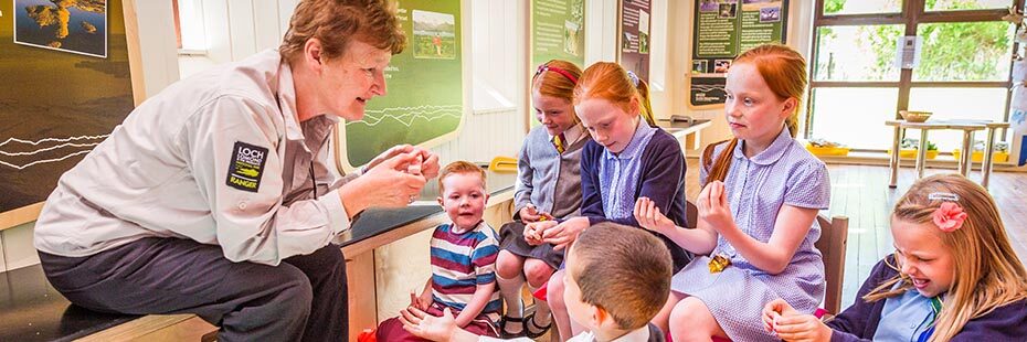 elderly-ranger-lady-with-national-park-uniform-shirt-chatting-to-group-of-six-young-children-in-classroom-area-of-balmaha-visitor-centre