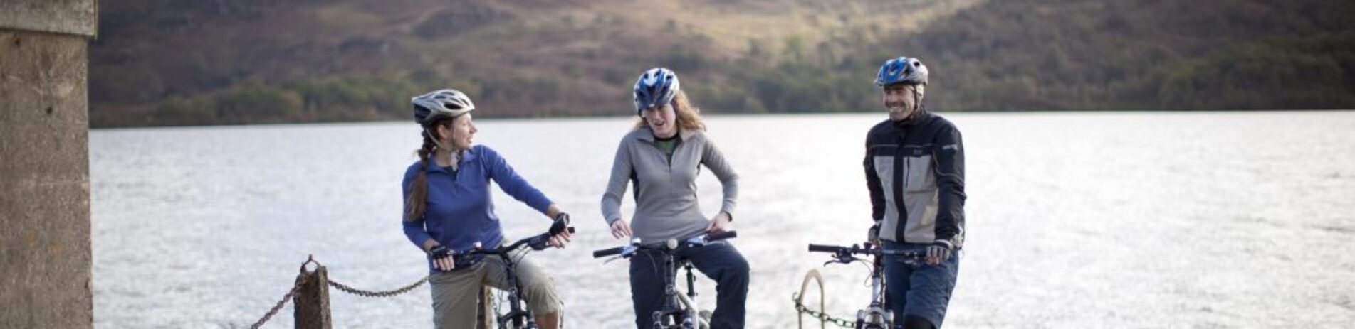 three-cyclists-on-bikes-with-helmets-on-at-edge-of-pier-with-loch-behind-them