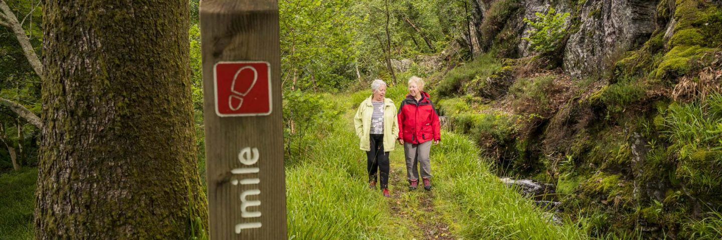 two-older-women-in-outdoor-gear-strolling-on-forest-path-near-ardentinny