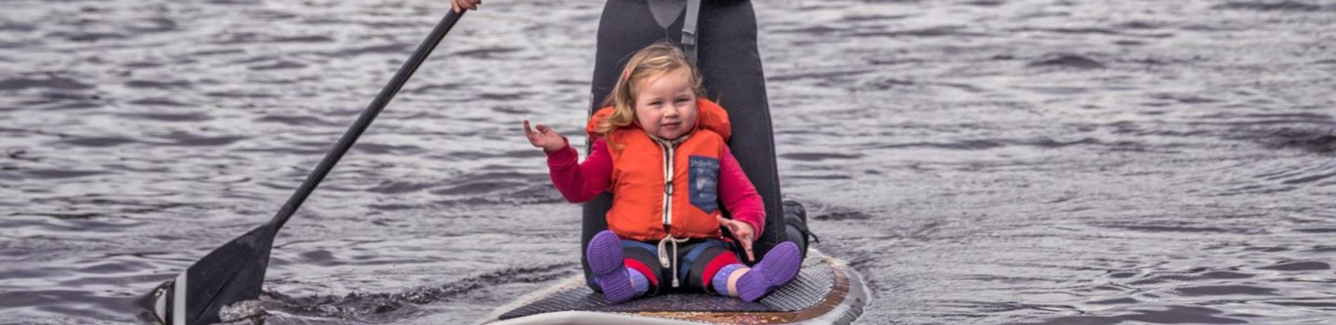 blonde-baby-girl-with-small-life-vest-sitting-on-paddleboard-and-waving-with-mums-legs-just-visible-behind-her-and-an-oar-on-the-left