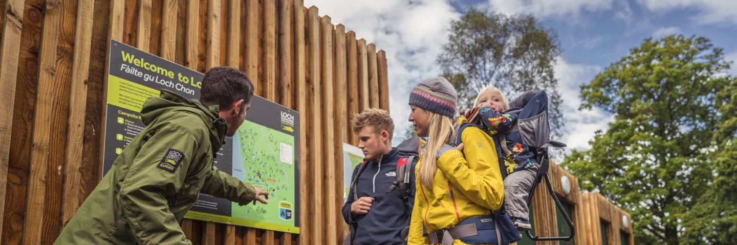 national-park-ranger-in-green-jacket-pointing-on-the-map-to-young-couple-with-baby-at-loch-chon-campsite