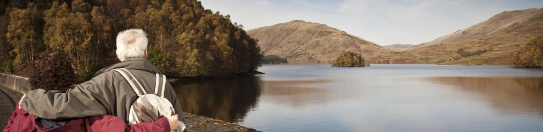 older-couple-embraced-affectionately-looking-into-distance-over-glen-finglas-reservoir-waters