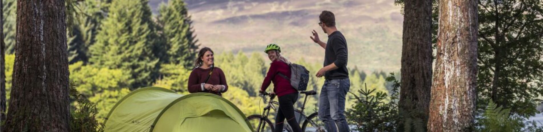young-couple-pitching-green-tent-at-three-lochs-forest-drive-wave-hello-to-passing-woman-cyclist