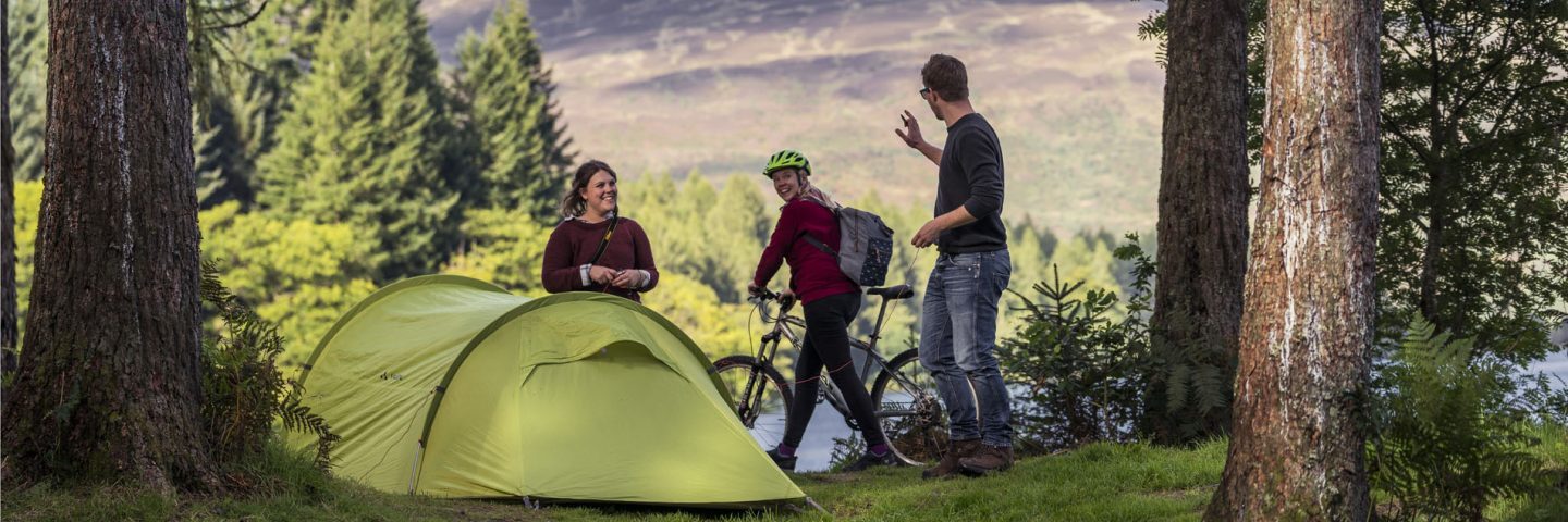 young-couple-pitching-green-tent-at-three-lochs-forest-drive-wave-hello-to-passing-woman-cyclist