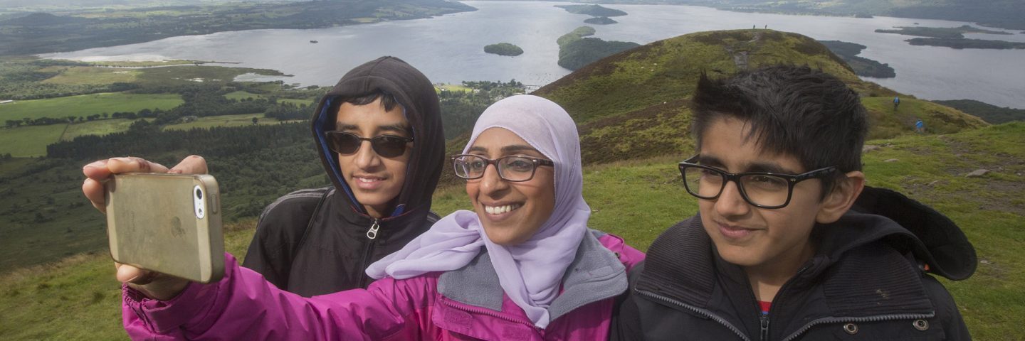 woman-wearing-glasses-and-hijab-taking-selfie-of-her-and-two-younger-boys-on-slopes-of-conic-hill-with-loch-lomond-and-islands-behind