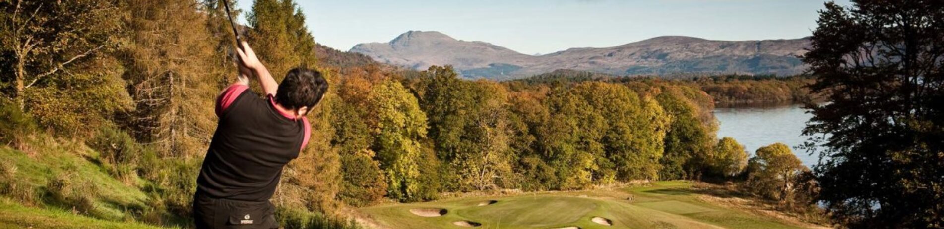 man-in-red-and-black-t-shirt-playing-gold-at-edge-of-forest-with-loch-lomond-visible-in-the-distance