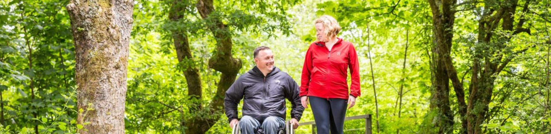 woman-in-jogging-gear-walking-alongside-man-in-wheelchair-on-path-surrounded-by-trees-in-balloch-castle-country-aprk