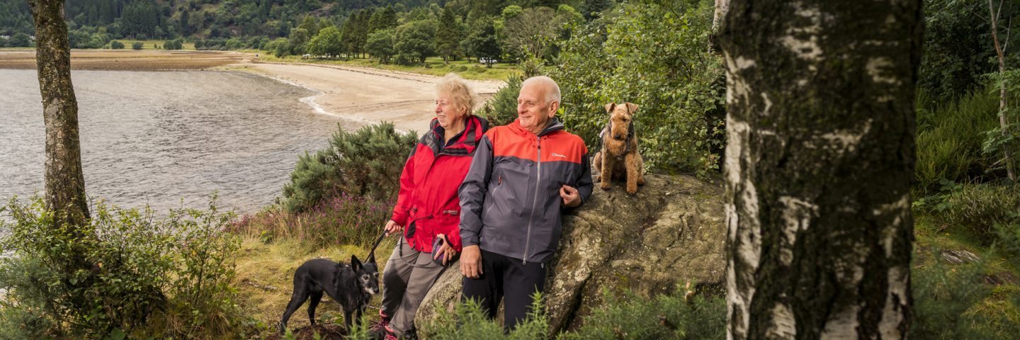 older-couple-with-two-dogs-looking-into-distance-by-large-rock-ardentinny-beach-and-forest-behind-them
