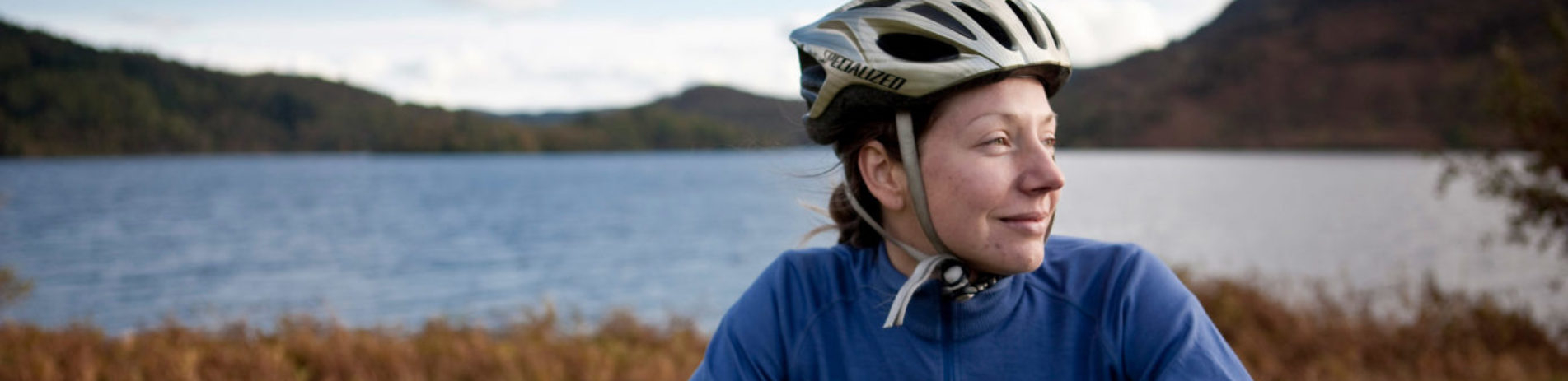 woman-in-blue-sweatshirt-and-bike-helmet-on-smiling-and-looking-in-the-distance-with-loch-and-hills-in-the-background