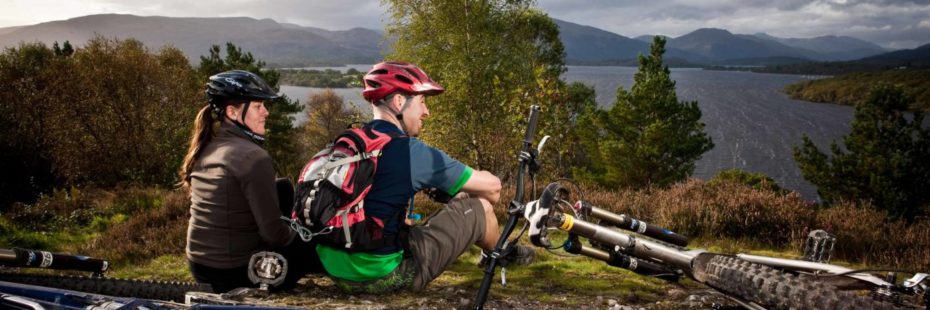 two-men-in-biking-gear-with-helmets-on-next-to-their-bikes-staring-in-the-distance-over-loch-lomond