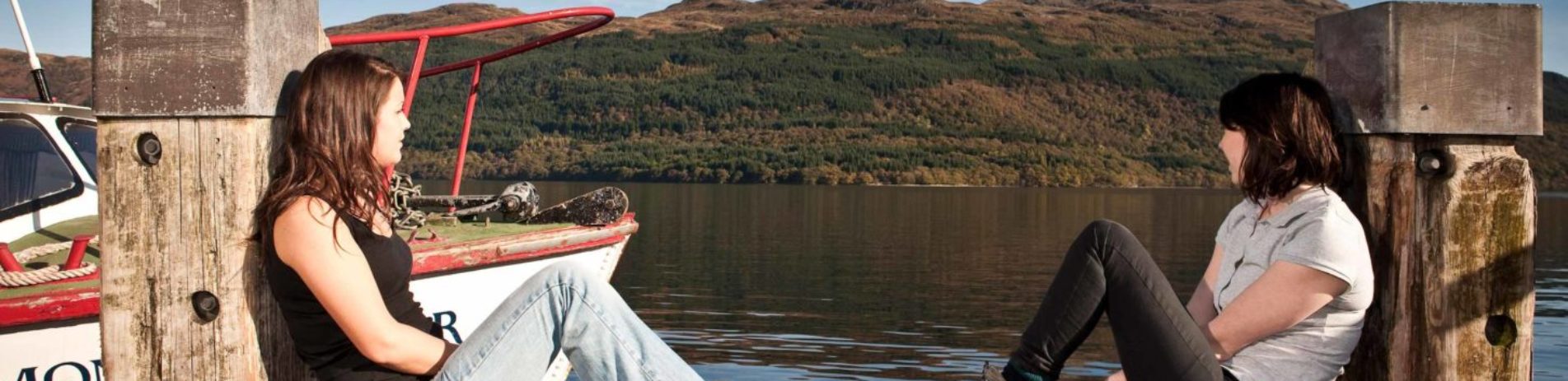 two-women-in-jeans-and-t-shirts-sitting-casually-at-edge-of-tarbet-pier-next-to-boat-looking-at-loch-lomond-water