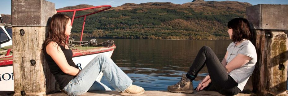 two-women-in-jeans-and-t-shirts-sitting-casually-at-edge-of-tarbet-pier-next-to-boat-looking-at-loch-lomond-water