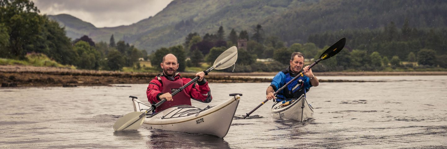 two-men-one-in-red-the-other-in-blue-peddling-separate-kayaks-on-loch-long-ardentinny-village-in-the-distance