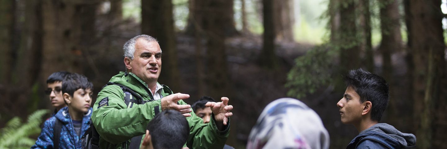 national-park-ranger-in-green-jacket-demonstrating-something-using-his-hands-in-front-of-school-students-in-the-forest