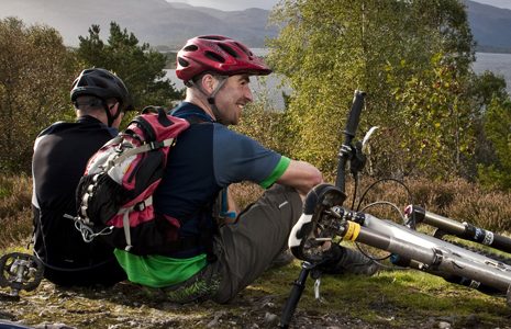two-cyclists-taking-a-rest-sitting-down-next-to-their-bikes-helmets-on-loch-lomond-visible-in-the-distance