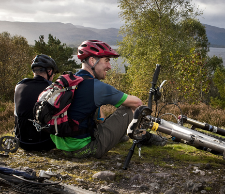 two-cyclists-taking-a-rest-sitting-down-next-to-their-bikes-helmets-on-loch-lomond-visible-in-the-distance
