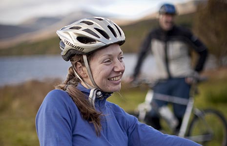 couple-with-bikes-and-wearing-bike-helmets-at-the-edge-of-loch-with-hills-in-distance