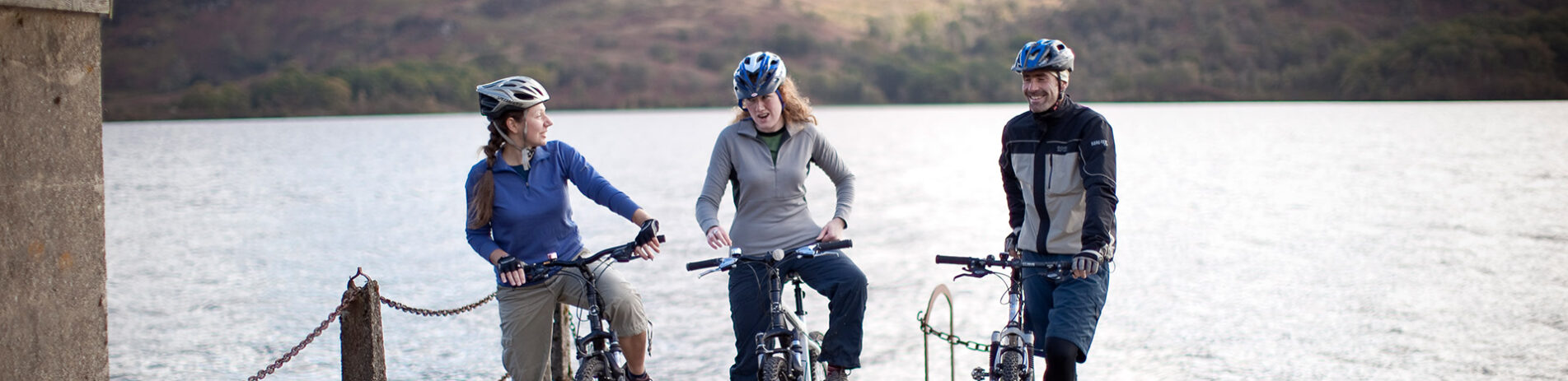 two-women-and-man-on-bikes-with-helmets-on-at-the-edge-of-pier-with-loch-behind-them