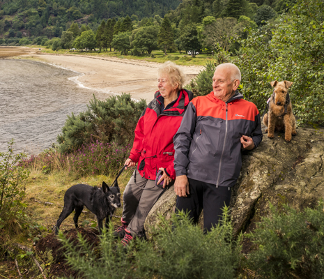 older-couple-with-two-dogs-looking-into-distance-by-large-rock-ardentinny-beach-and-forest-behind-them