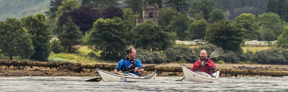 two-men-one-in-red-the-other-in-blue-peddling-separate-kayaks-on-loch-long-ardentinny-village-in-the-distance