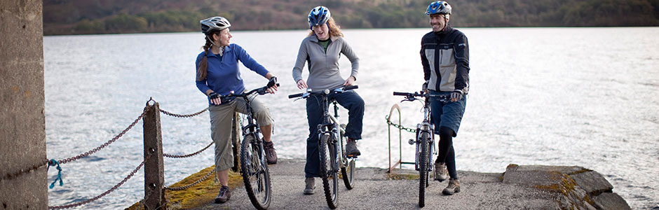 two-women-and-man-on-bikes-with-helmets-on-at-the-edge-of-pier-with-loch-behind-them