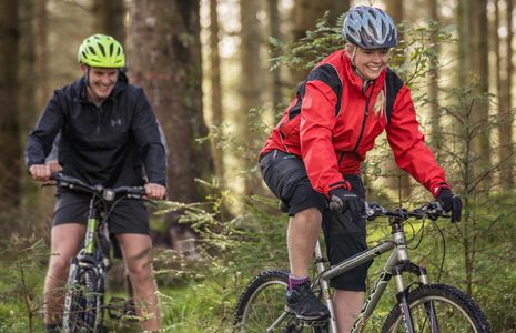 man-and-woman-in-outdoor-gear-cycling-on-forest-track-downhill-and-smiling