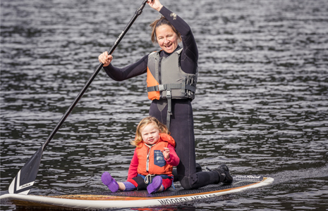 woman-in-wetsuit-and-vest-kneeling-on-paddleboard-holding-oar-and-smiling-with-very-young-daughter-at-her-feet-looking-at-camera-surrounded-by-waters