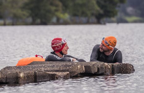 woman-and-man-in-wetsuits-and-swim-caps-chatting-next-to-pier-while-half-submerged-in-water