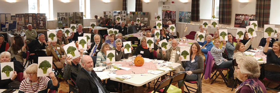 local-village-community-gathering-in-village-hall-of-dozens-of-people-all-holding-small-boards-with-broccoli-plant-illustrated-on-them
