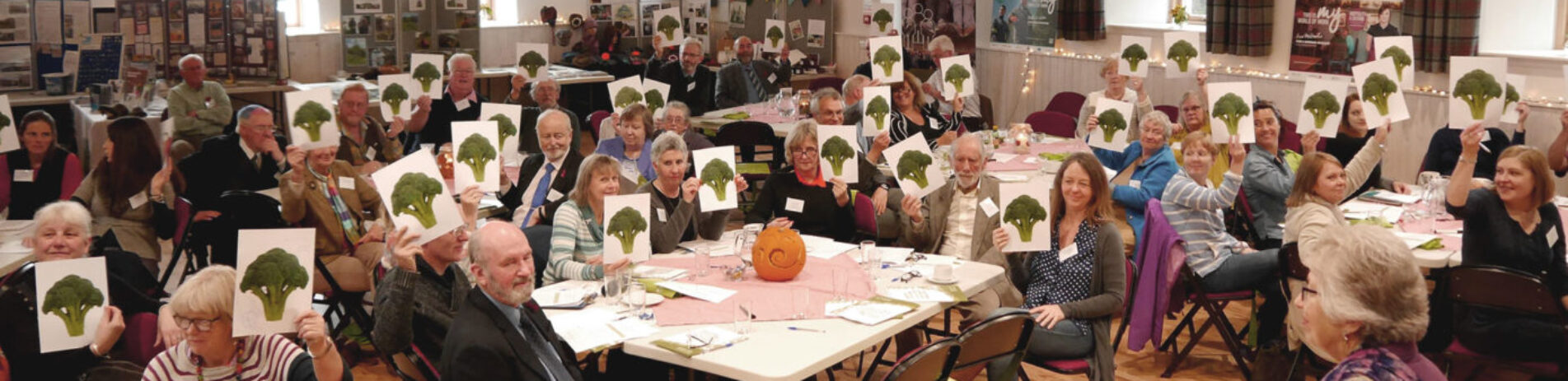 local-village-community-gathering-in-village-hall-of-dozens-of-people-all-holding-small-boards-with-broccoli-plant-illustrated-on-them