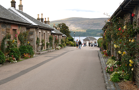 luss-village-pier-road-with-loch-lomond-and-tourists-in-the-distance-and-stone-houses-decorated-with-flowers-on-both-sides-of-the-road