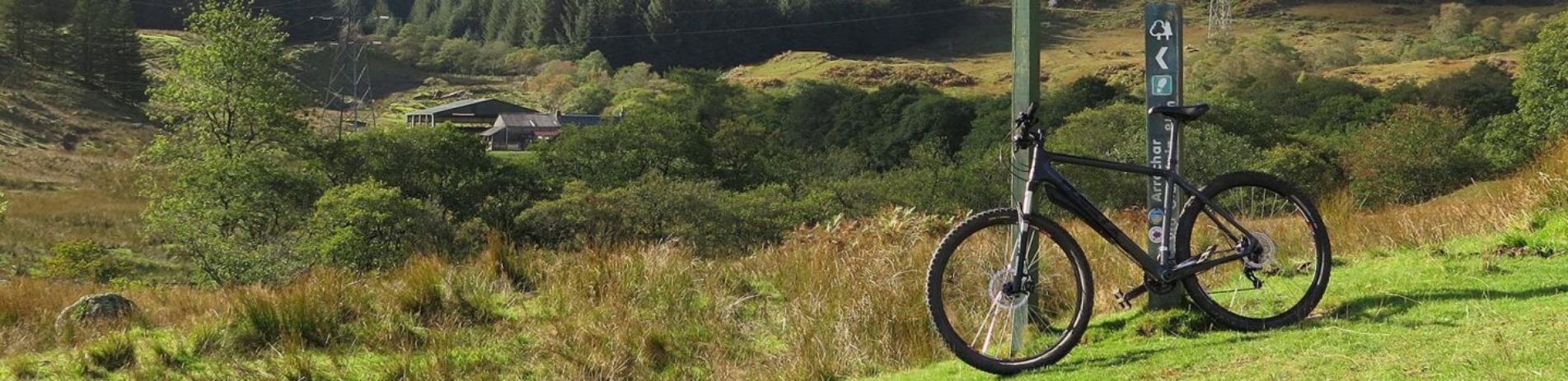 bike-propped-against-direction-wooden-pole-above-forested-landscape-and-next-to-track-in-glen-loin