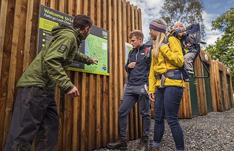 national-park-ranger-in-branded-dark-green-jacket-showing-young-blond-couple-with-baby-something-on-campsite-map-at-loch-chon-chon-toilet-blocks-with-green-doors-behind