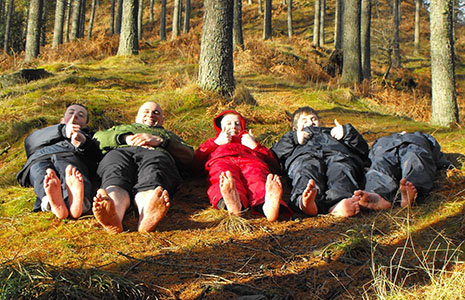 five-children-and-ranger-all-dressed-in-waterproofs-lying-on-forest-floor-barefoot-with-thumbs-up