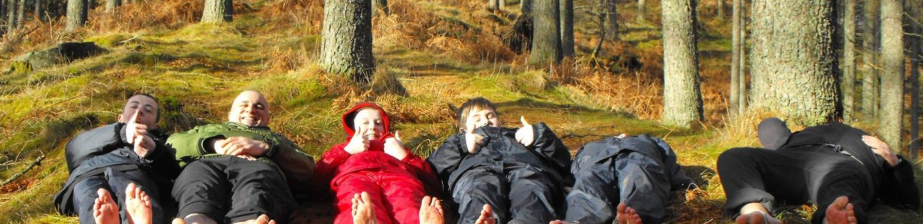 five-children-and-ranger-all-dressed-in-waterproofs-lying-on-forest-floor-barefoot-with-thumbs-up