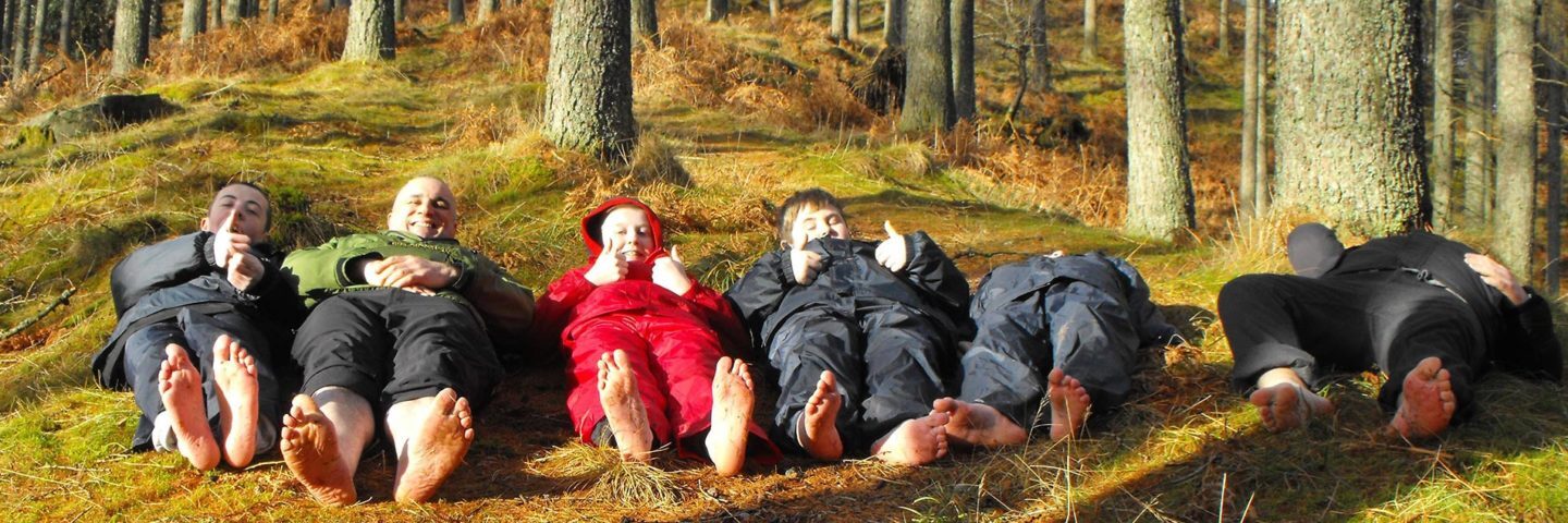 five-children-and-ranger-all-dressed-in-waterproofs-lying-on-forest-floor-barefoot-with-thumbs-up
