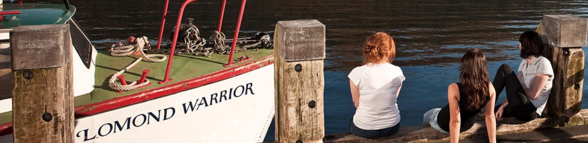 group-of-three-young-women-at-edge-of-tarbet-pier-looking-at-the-water-on-loch-lomond-next-to-moored-boat