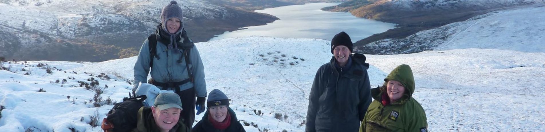 group-of-volunteers-wrapped-up-warm-in-winter-jackets-with-dog-on-snowy-hill-with-loch-arklet-and-mountains-behind