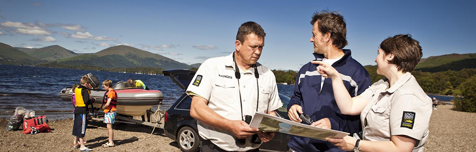 two-rangers-chatting-to-member-of-public-and-pointing-on-map-and-in-the-distance-with-boat-being-launched-in-loch-lomond-next-to-car-and-group-of-children