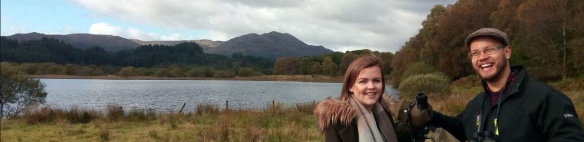 two-young-people-girl-and-boy-next-to-camera-tripod-boy-is-holding-binoculars-at-edge-of-loch-venachar-ben-venue-visible-in-the-distance-bird-watching