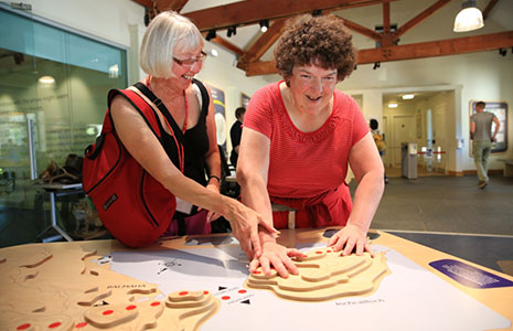 blind-woman-in-red-dress-with-curly-brown-hair-being-assisted-by-blonde-woman-in-touching-wooden-contours-of-highland-boundary-line-terrain-map-in-balmaha-visitor-centre