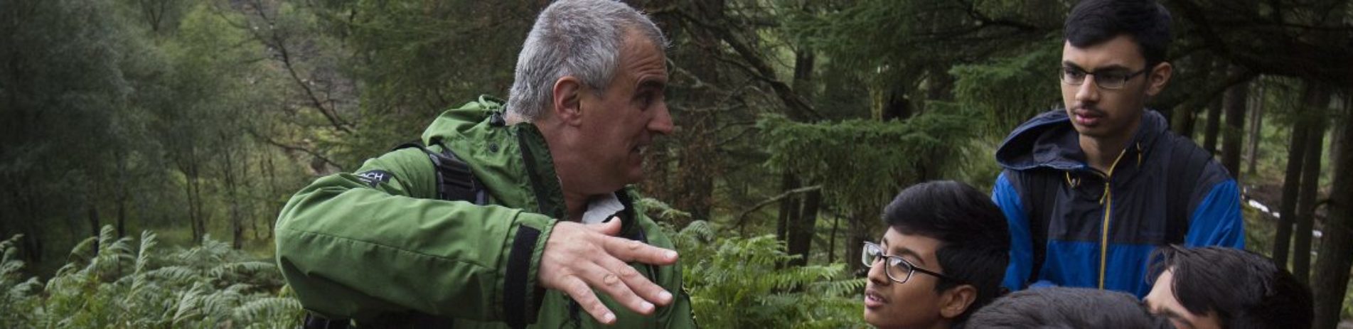 national-park-ranger-in-green-jacket-demonstrating-something-using-his-hands-in-front-of-school-students-in-the-forest